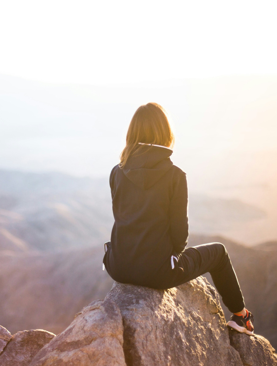 Ansicht einer Frau von hinten, in Kapuzenjacke und Sportschuhen, die in der Morgensonne auf einem Felsen sitzt und an einem sehr hohen Punkt in die Weite schaut, im Hintergrund unscharf Berge und Horizont