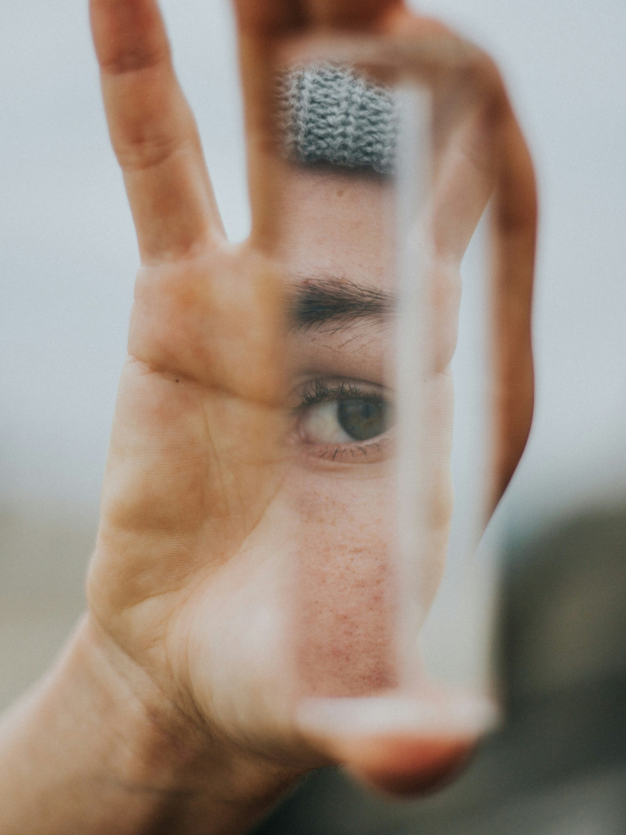 Eine Handinnenfläche hält ein Hochkant-Glas-Objekt zwischen Daumen und Zeigefinger, in dem sich ein Auge sowie der schmale Ausschnitt eines Gesichts zeigt
