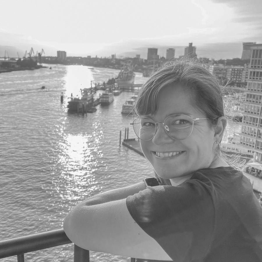 Christiane Häßler, schwarz-weiß Bild, auf ein hohes Balkongeländer gelehnt vor einem großen Fluß, seitlich über die linke Schulter schauend mit freundlichem Lächeln