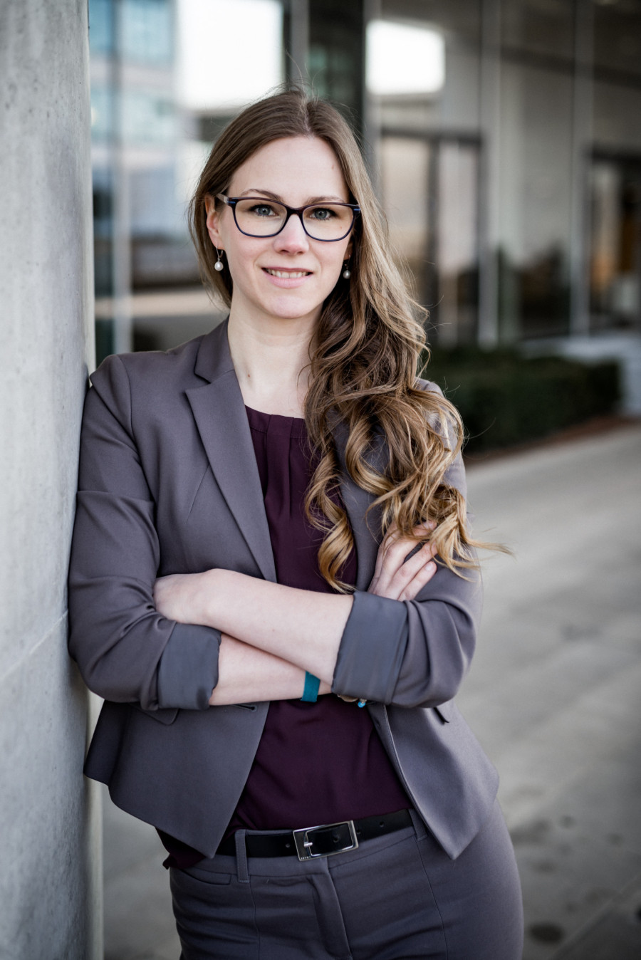 Melanie Eberle, im Kostüm mit Arme hochgekrämpelt, Arme lässig verschränkt, draußen mit Gebäude im Hintergrund an eine Wand angelehnt, freundlichen Lächeln