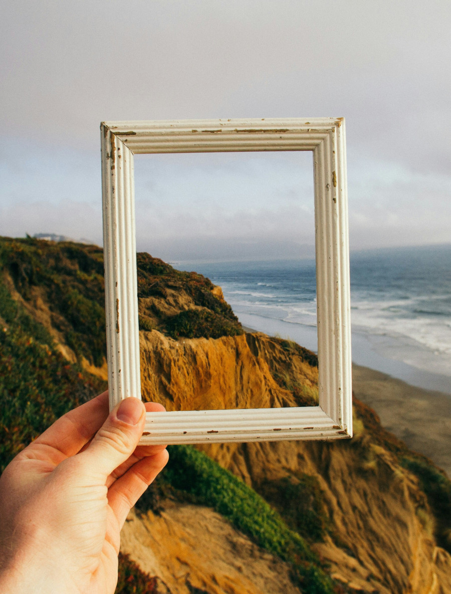 Eine Hand hält einen leeren Bilderrahmen vor die Kulisse einer hohen Küstenlandschaft, mit Meer und Strand, und verhangenem Himmel im Hintergrund